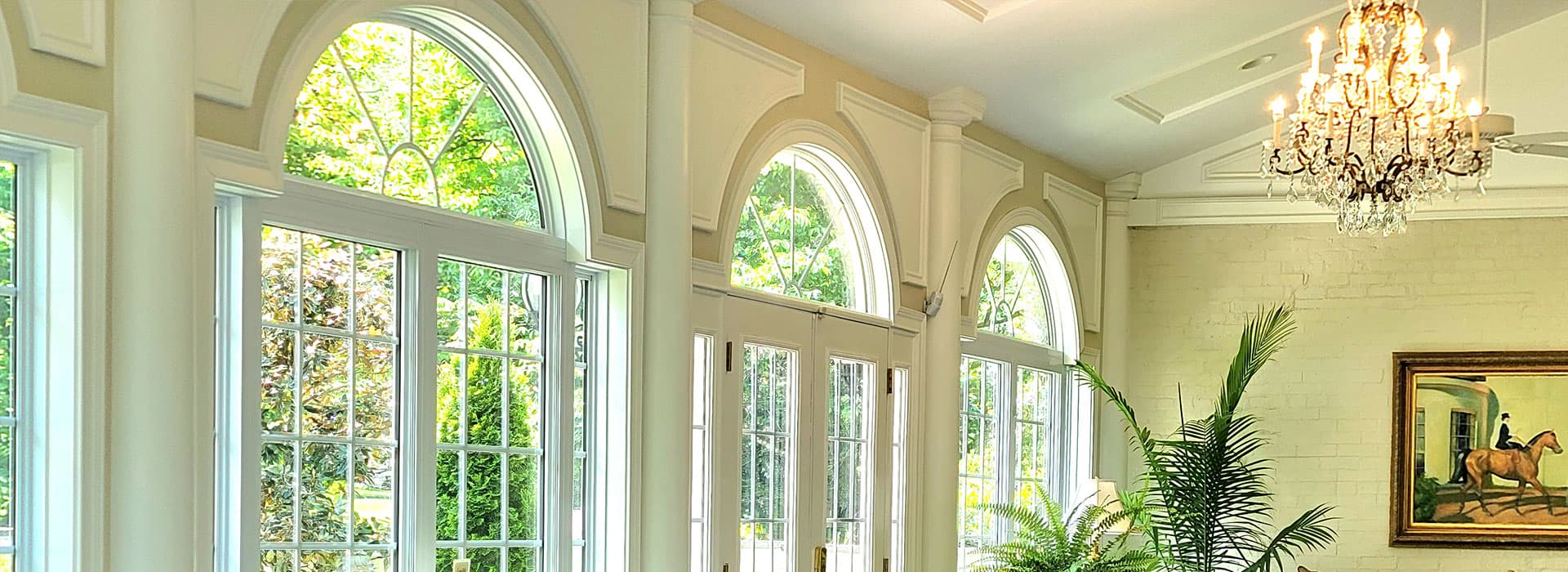 Bright sunlit room with large arched windows, greenery, and a chandelier.