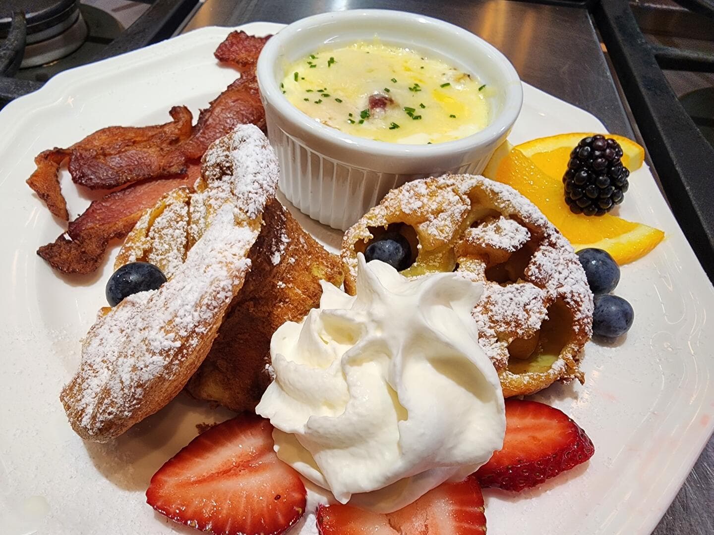 A plate of breakfast featuring crispy bacon, a creamy dish, and a variety of sweet pastries, fresh fruits, and whipped cream.